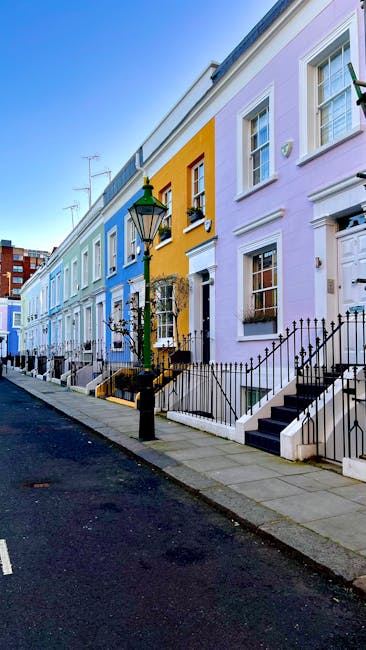 A row of colorful pastel terraced houses along a quiet street in Dulwich, featuring front gardens enclosed by black metal railings. The houses are painted in shades of light blue, yellow, lavender, an
