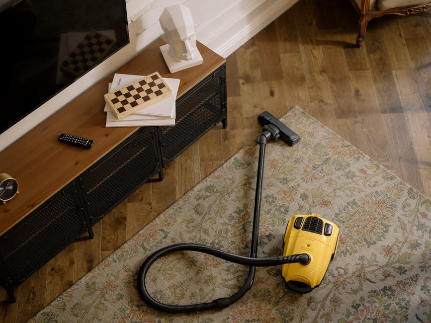 A woman with brown hair tied back, wearing a light-striped shirt and blue jeans, is kneeling on a patterned rug in a modern living room, cleaning a wooden floor with a blue cloth. The room features a 
